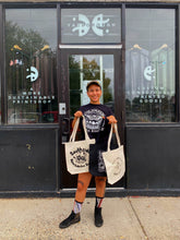 Load image into Gallery viewer, Alex is holding three of the tote bags in front of Yarborough Print Shop. She is also wearing the Butterfly shirt in a size medium. 
