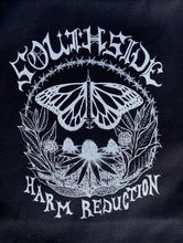 Load image into Gallery viewer, The Butterfly Tote has a butterfly in the center of the image whos body is a needle. There are flowers below the butterfly forming a half circle. The top part of the circle is formed by barbed wire. The words &quot;Southside&quot; are above the circle and &quot;Harm Reduction&quot; are below the circle. Image is on the black tote. 
