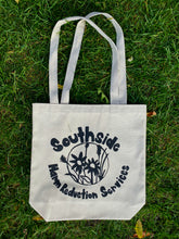 Load image into Gallery viewer, The Circle Tote has a circle in the center of the bag with flowers in the center. The word &quot;Southside&quot; is above the circle and &quot;Harm Reduction Services&quot; is below the circle. Image is on the natural tote. 
