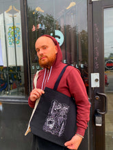 Load image into Gallery viewer, Jake is holding the Safe Use Supplies tote in front of Yarborough Print Shop. He is looking at the camera and wearing maroon hoodie. 
