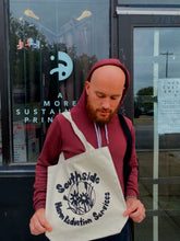 Load image into Gallery viewer, Jake is holding the Circle tote in front of Yarborough Print shop. He is wearing a maroon hoodie and looking down at the bag.
